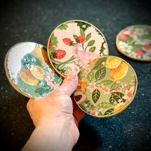 May include: Four round coasters with gold trim and floral designs. The coasters feature different floral patterns, including lemons, strawberries, and flowers.