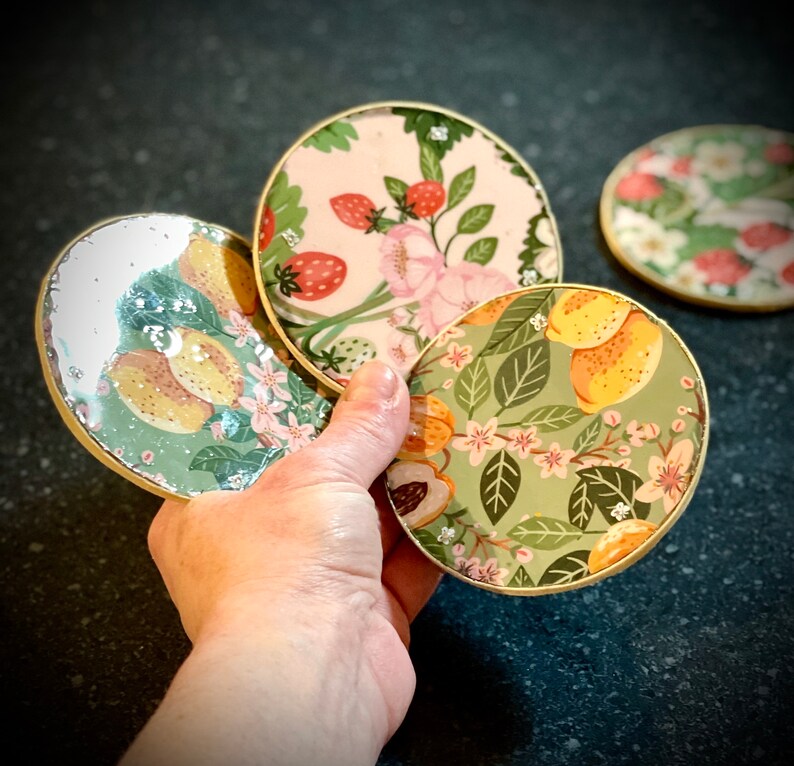 May include: Four round coasters with gold trim and floral designs. The coasters feature different floral patterns, including lemons, strawberries, and flowers.