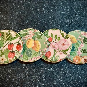 May include: Four round green and white coasters with floral and fruit designs. The coasters feature strawberries, lemons, peaches, and flowers.
