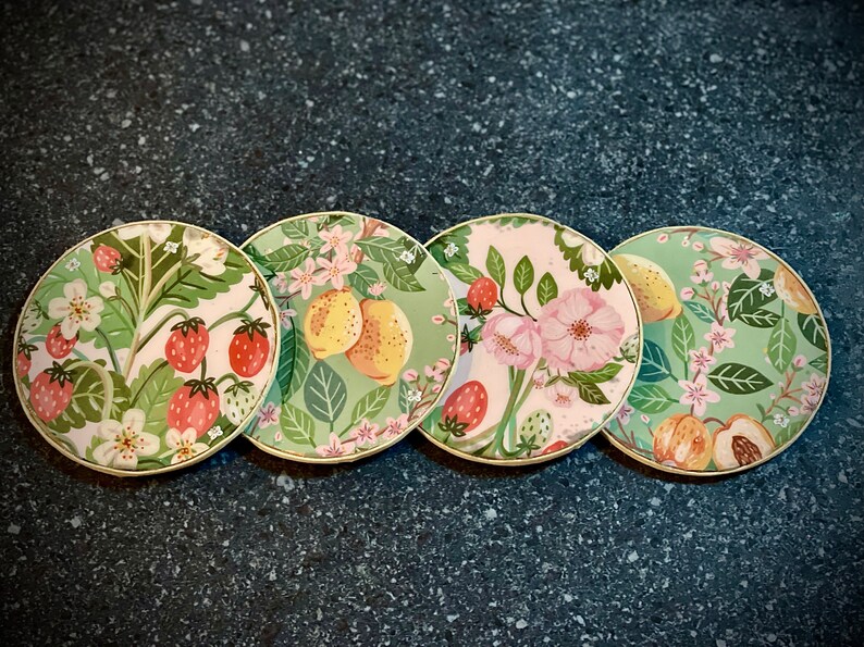 May include: Four round green and white coasters with floral and fruit designs. The coasters feature strawberries, lemons, peaches, and flowers.