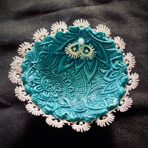 May include: A teal blue ceramic dish with intricate floral designs, surrounded by a white lace trim. The dish has a small white bead in the centre.