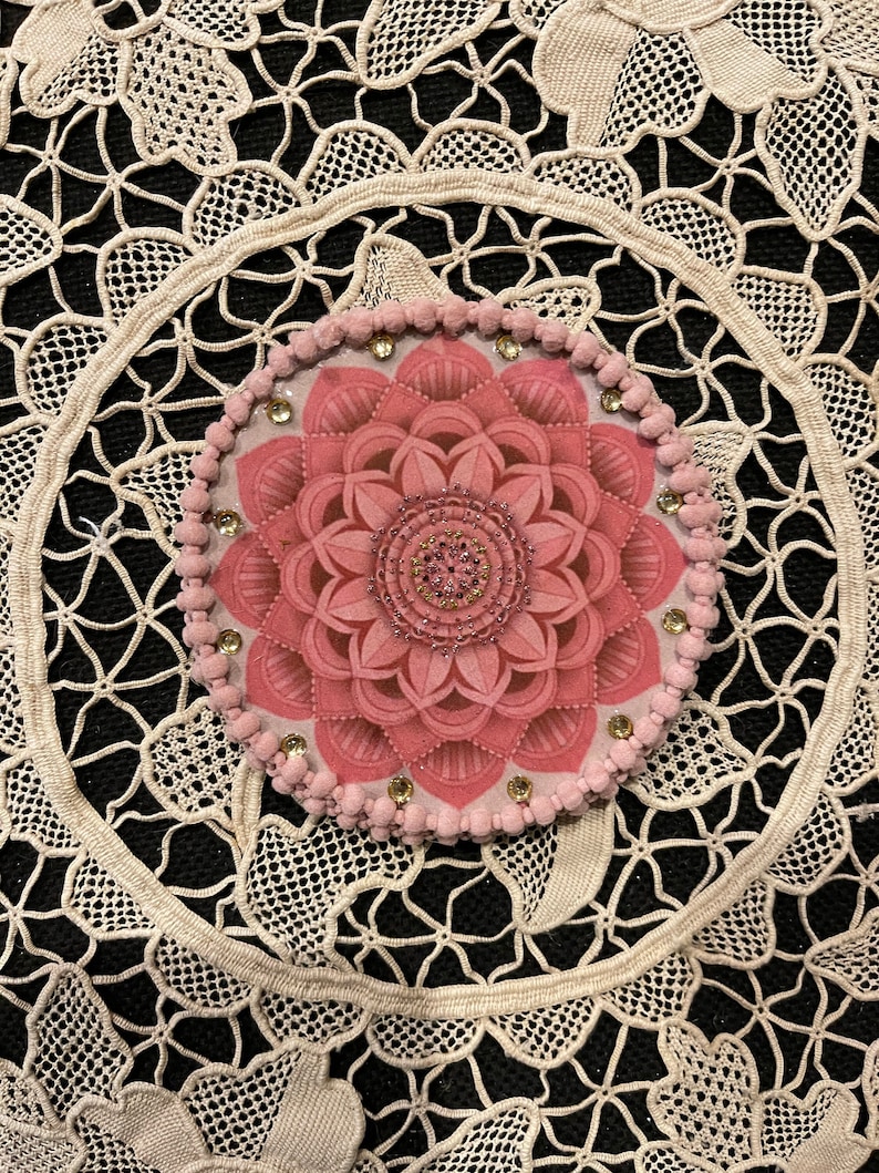 K&ouml;nnte beinhalten: Ein rosa Blumendesign, m&ouml;glicherweise ein Untersetzer oder Dekorationselement, ist auf einer schwarzen Oberfl&auml;che zentriert. Das Design zeigt eine detaillierte, geschichtete Blume mit einem Perlenmittelpunkt und ist von einem Ring aus kleinen rosa Pompons und Strasssteinen umgeben. Der Hintergrund ist eine cremefarbene Spitzendecke.