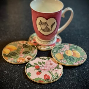 May include: Four round coasters with floral designs. The coasters have a gold rim and feature different floral patterns, including strawberries, peaches, and lemons. A pink mug with a cat illustration and a heart design sits on top of one of the coasters.