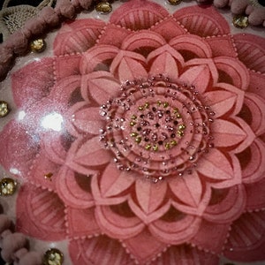 K&ouml;nnte beinhalten: Nahaufnahme eines dekorativen, rosa Blumendesigns mit einem verzierten Mittelkreis. Die Bl&uuml;tenbl&auml;tter sind geschichtet und strukturiert. Das Design ist mit kleinen, funkelnden Edelsteinen und einer Bord&uuml;re aus rosa Pompons und goldfarbenen Perlen akzentuiert.