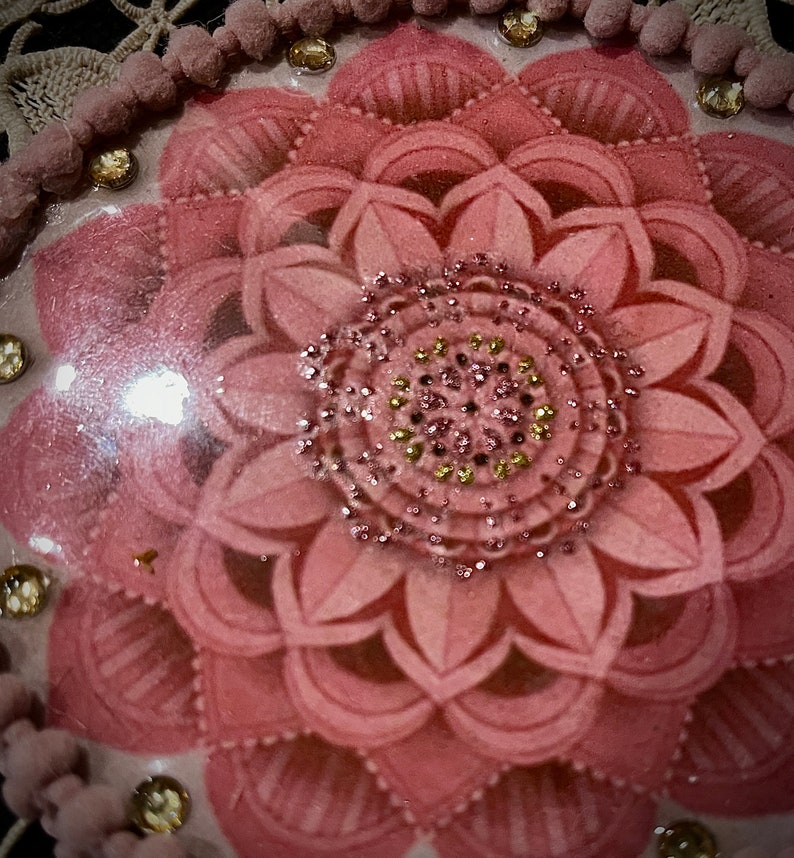 K&ouml;nnte beinhalten: Nahaufnahme eines dekorativen, rosa Blumendesigns mit einem verzierten Mittelkreis. Die Bl&uuml;tenbl&auml;tter sind geschichtet und strukturiert. Das Design ist mit kleinen, funkelnden Edelsteinen und einer Bord&uuml;re aus rosa Pompons und goldfarbenen Perlen akzentuiert.