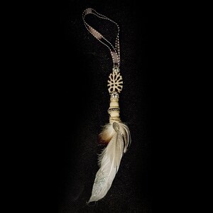 Puede incluir: Un adorno colgante decorativo que presenta un lazo de cinta marrón, una cuenta ovalada tallada de color crema con un patrón floral, espaciadores plateados con cristales transparentes y un grupo de plumas naturales con purpurina blanca.