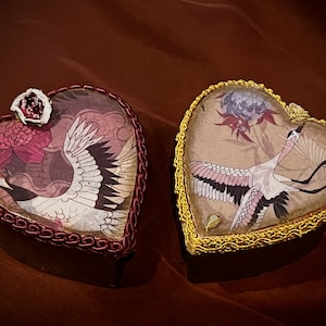 Puede incluir: Dos cajas decorativas en forma de corazón. Una caja tiene un borde rojo y una ilustración de grulla con una flor rosa. La otra caja tiene un borde dorado y una ilustración de grulla con una flor morada. Ambas cajas tienen una estética vintage.
