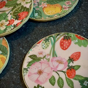 May include: Four round coasters with a gold rim. Each coaster features a different floral design with pink flowers, green leaves, and red strawberries. One coaster has a green background with yellow lemons.