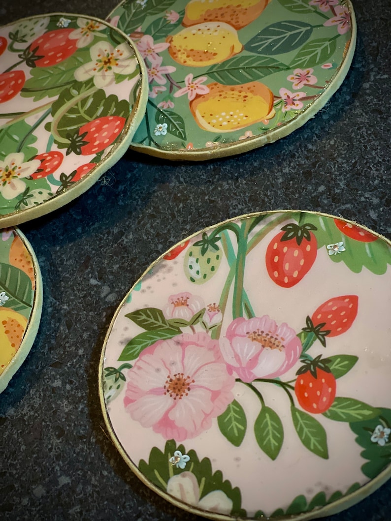 May include: Four round coasters with a gold rim. Each coaster features a different floral design with pink flowers, green leaves, and red strawberries. One coaster has a green background with yellow lemons.