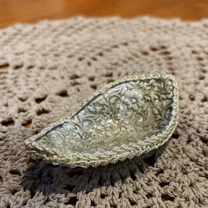 May include: A small, silver-coloured, decorative bowl with a beaded rim. The bowl has a floral design and is sitting on a crocheted surface.