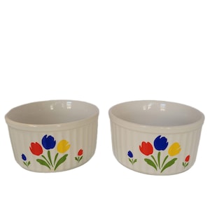 Puede incluir: Dos ramequines de cerámica blanquecina con un diseño de tulipanes rojos, azules y amarillos. Los ramequines tienen un exterior acanalado y un interior liso. El diseño floral está pintado en la parte delantera de cada ramequín.