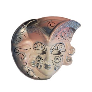 May include: A decorative mask with a winking face and a smiling face. The mask is painted in shades of pink, silver, and black, with glitter accents. The mask has a curved shape and is likely for a masquerade or costume event.