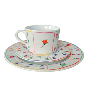 Puede incluir: Un juego de taza y platillo de porcelana blanca con un diseño floral colorido. La taza y el platillo presentan un patrón repetitivo de pequeñas flores con contornos coloridos. La taza tiene una sola flor más grande con un centro rojo.
