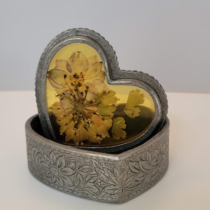 Peut inclure: Sous-verre en forme de cœur avec une fleur jaune pressée, enfermée dans de la résine transparente, dans un support argenté décoratif. Le support présente un motif floral en relief. Un objet de décoration unique.