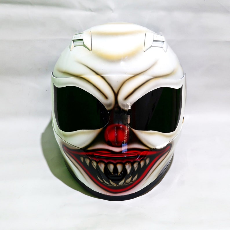 May include: A white motorcycle helmet with a painted design of a clown face with a red nose and a wide, toothy grin.