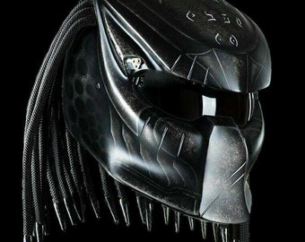 May include: A black motorcycle helmet with a Predator-inspired design. The helmet has a black and gray camouflage pattern and a black visor. The helmet has a long, black braid hanging from the back.