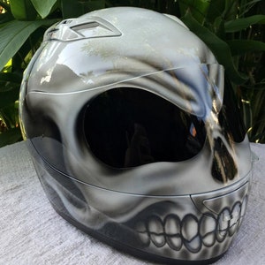 Puede incluir: Un casco de moto blanco con un dise&ntilde;o de calavera pintado en &eacute;l. El casco tiene una visera negra y una correa de barbilla negra.