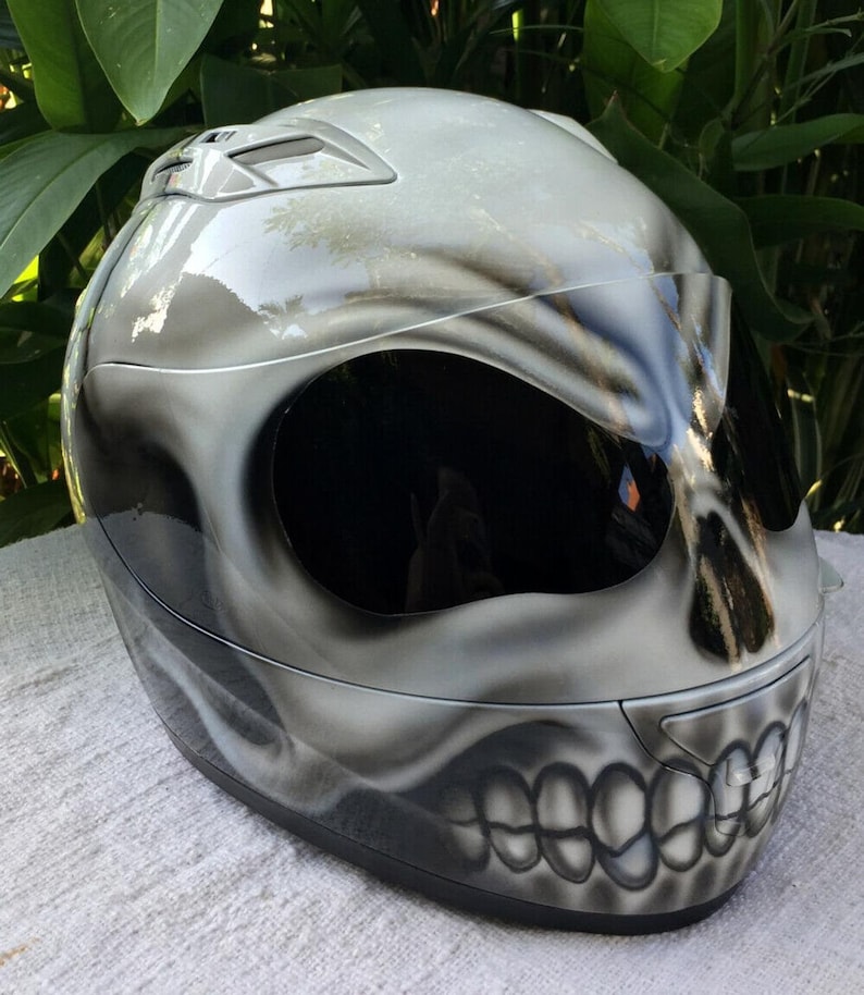 Puede incluir: Un casco de moto blanco con un dise&ntilde;o de calavera pintado en &eacute;l. El casco tiene una visera negra y una correa de barbilla negra.
