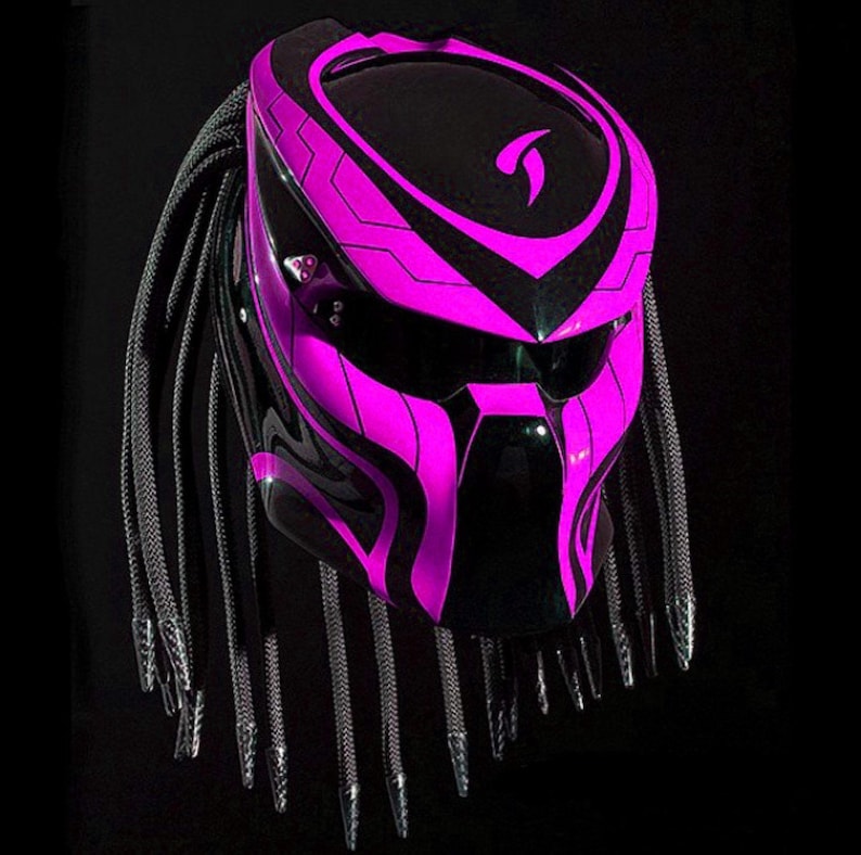 K&ouml;nnte beinhalten: Ein schwarzer und pinkfarbener Motorradhelm mit einem Predator-Design und schwarzen Dreadlocks. Der Helm hat ein schwarzes Visier und ein pinkfarbenes und schwarzes Design auf der Vorderseite.