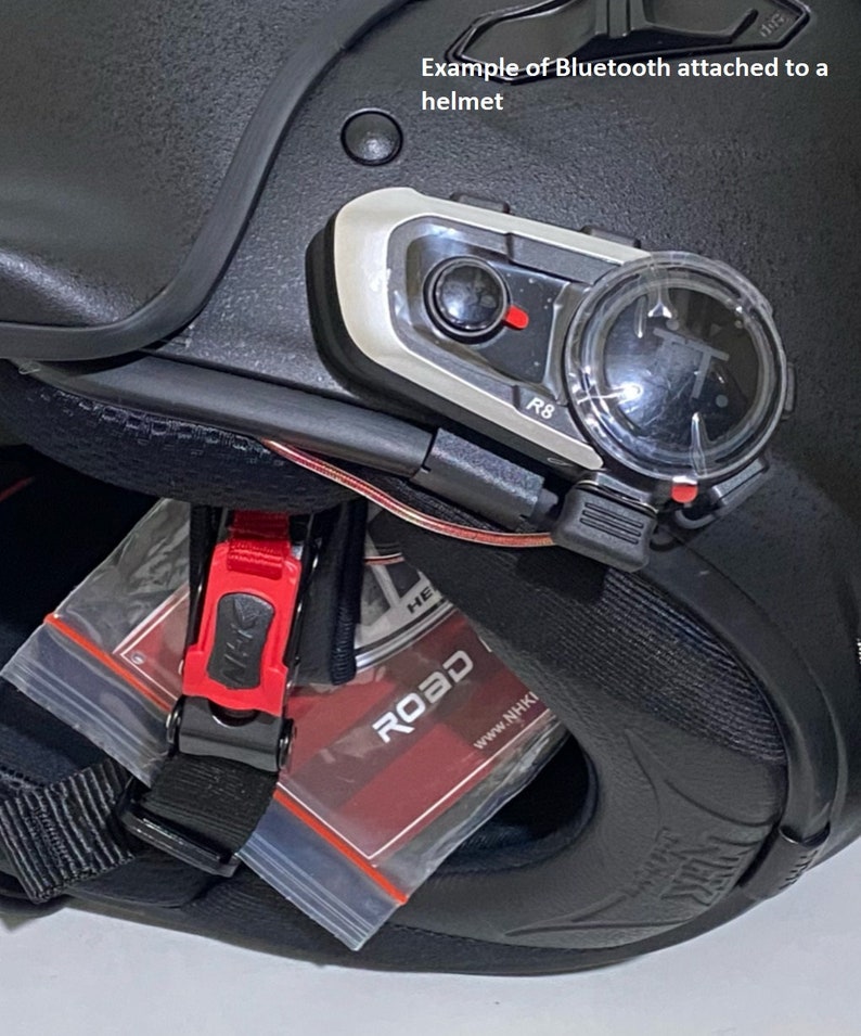 K&ouml;nnte beinhalten: Nahaufnahme eines schwarzen Motorradhelms mit einem silbernen und schwarzen Bluetooth-Kommunikationsger&auml;t, das an der Seite befestigt ist. Das Ger&auml;t hat einen roten Knopf und einen kleinen Bildschirm. Der Helm hat einen schwarzen Riemen und eine rote Schnalle. Der Text "ROAD" ist auf einem roten Etikett sichtbar.