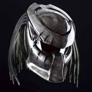 May include: A silver motorcycle helmet with a black and green braided strap. The helmet has a predator-like design with a textured surface and a dark visor.