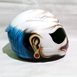 May include: A white motorcycle helmet with a blue and yellow clown face design. The helmet has a black visor and a red nose.