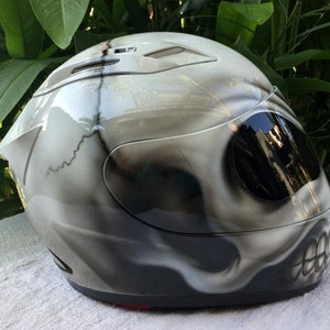 Puede incluir: Un casco de moto blanco con un dise&ntilde;o de calavera pintado en &eacute;l. El casco tiene una visera oscura tintada y una correa de barbilla negra.