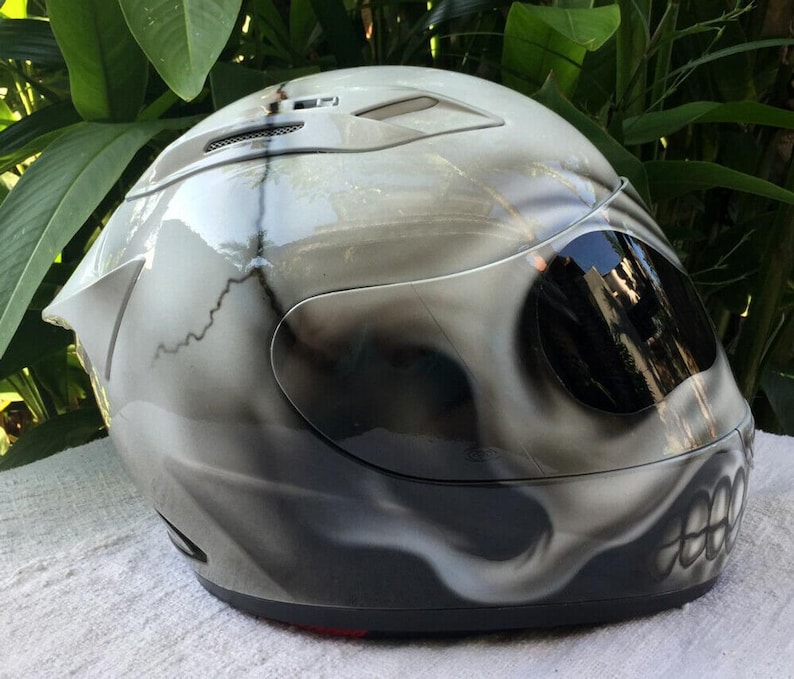 Puede incluir: Un casco de moto blanco con un dise&ntilde;o de calavera pintado en &eacute;l. El casco tiene una visera oscura tintada y una correa de barbilla negra.