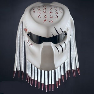 May include: White motorcycle helmet with a Predator design. The helmet has a black visor, white dreadlocks, and red markings. The helmet has a black and white design on the front with the word "J.C.M." written on the side.