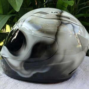 Puede incluir: Un casco de moto blanco con un dise&ntilde;o de calavera en blanco y negro. El casco tiene una visera transparente y una correa de barbilla negra.