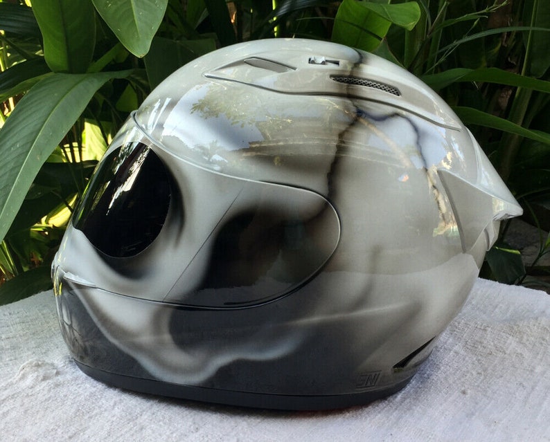 Puede incluir: Un casco de moto blanco con un dise&ntilde;o de calavera en blanco y negro. El casco tiene una visera transparente y una correa de barbilla negra.