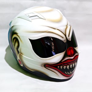 May include: White motorcycle helmet with a clown face painted on it. The clown has a red nose, a wide smile with sharp teeth, and black eyes.