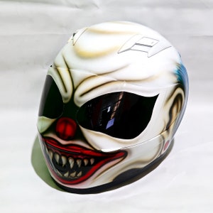 May include: A white motorcycle helmet with a clown face painted on it. The clown has a red nose, red lips, and white teeth. The helmet has a black visor.