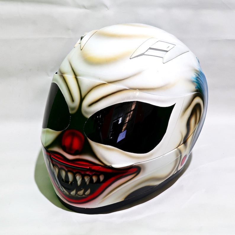 May include: A white motorcycle helmet with a clown face painted on it. The clown has a red nose, red lips, and white teeth. The helmet has a black visor.