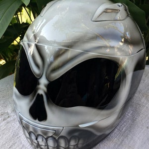 Puede incluir: Un casco de moto blanco con un dise&ntilde;o de calavera pintado en la parte delantera. El casco tiene una visera negra y un fondo blanco con detalles grises.