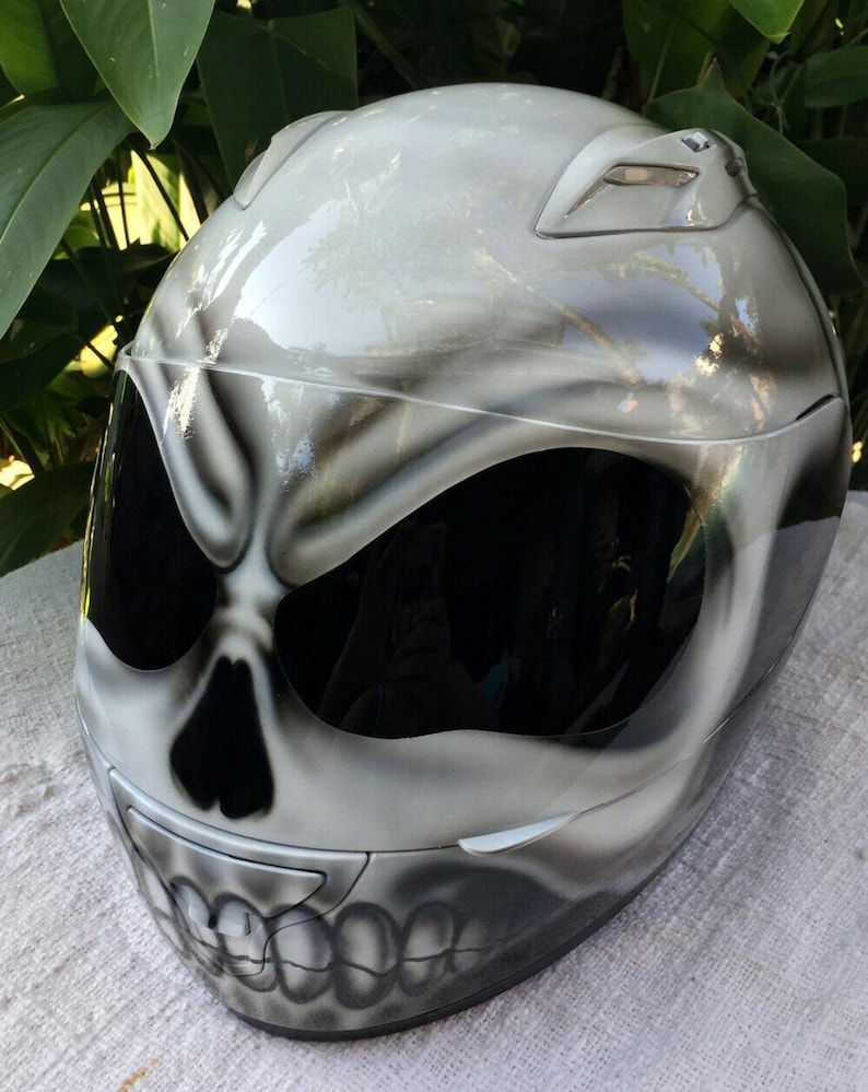 Puede incluir: Un casco de moto blanco con un dise&ntilde;o de calavera pintado en la parte delantera. El casco tiene una visera negra y un fondo blanco con detalles grises.