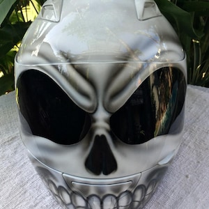 Puede incluir: Un casco de moto blanco con un dise&ntilde;o de calavera pintado en la parte delantera. La calavera tiene ojos negros y una boca negra con dientes.