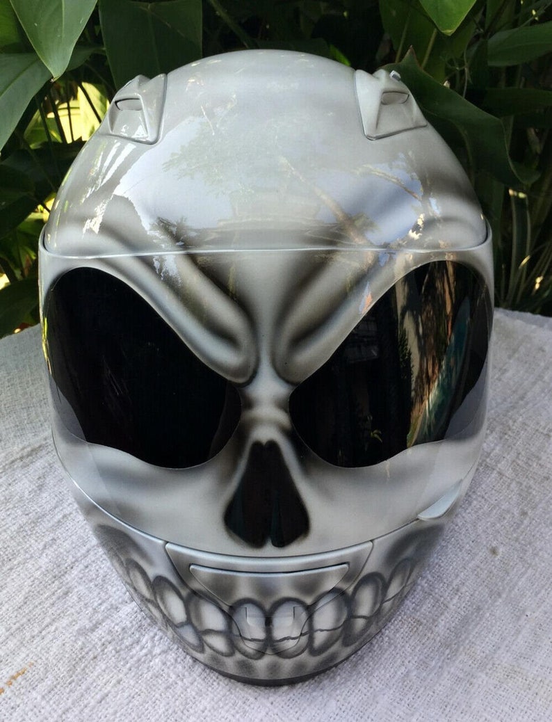 Puede incluir: Un casco de moto blanco con un dise&ntilde;o de calavera pintado en la parte delantera. La calavera tiene ojos negros y una boca negra con dientes.