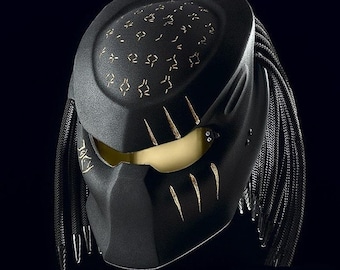 May include: A black motorcycle helmet with a gold design and a black braided strap. The helmet has a predator-like design with a visor and a textured surface.