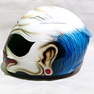 May include: White motorcycle helmet with a blue and white clown face airbrushed design. The helmet has a black visor and a red nose.
