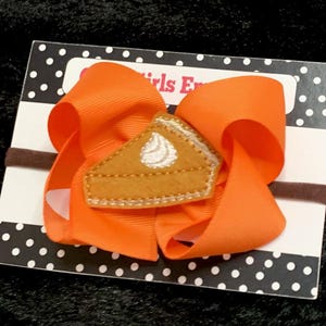 May include: Orange hair bow with a felt pumpkin pie applique. The bow is attached to a brown elastic headband.
