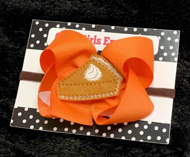 May include: Orange hair bow with a felt pumpkin pie applique. The bow is attached to a brown elastic headband.