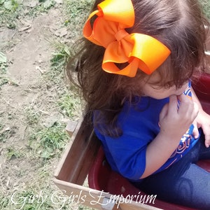 May include: A young child wearing a blue shirt with a white and orange design sits in a red wagon. The child has a large orange bow in their hair.