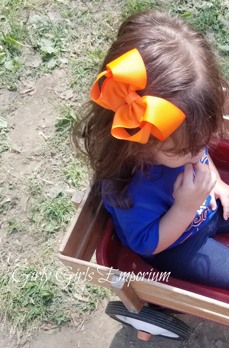 May include: A young child wearing a blue shirt with a white and orange design sits in a red wagon. The child has a large orange bow in their hair.