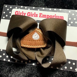 May include: A brown hair bow with a brown elastic headband. The bow has a felt pie slice with white frosting applique in the center. The card reads "Girly Girls Emporium".
