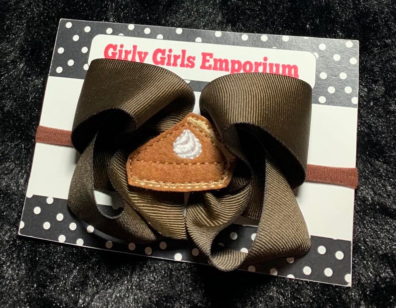 May include: A brown hair bow with a brown elastic headband. The bow has a felt pie slice with white frosting applique in the center. The card reads "Girly Girls Emporium".