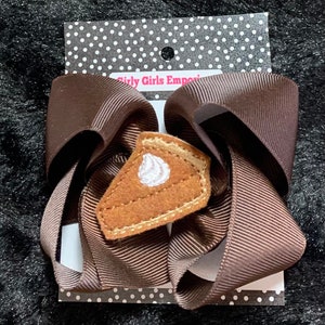 May include: A brown ribbon bow with a felt pumpkin pie applique. The pie has a white swirl of whipped cream on top. The bow is on a white card with black polka dots and the text "Girly Girls Emporium".