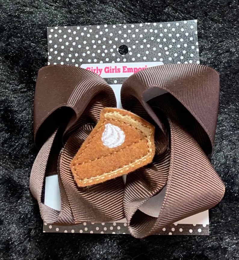 May include: A brown ribbon bow with a felt pumpkin pie applique. The pie has a white swirl of whipped cream on top. The bow is on a white card with black polka dots and the text "Girly Girls Emporium".