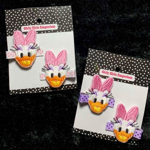 May include: Two pairs of hair clips featuring embroidered cartoon duck faces with pink bows. The clips are on white cards with black polka dots and the text "Girly Girls Emporium".