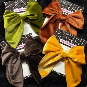 May include: Four velvet hair bows in shades of green, orange, brown, and yellow. Each bow is attached to a black and white polka dot card with the text "Girly Girls Emporium". The bows are arranged on a black surface.